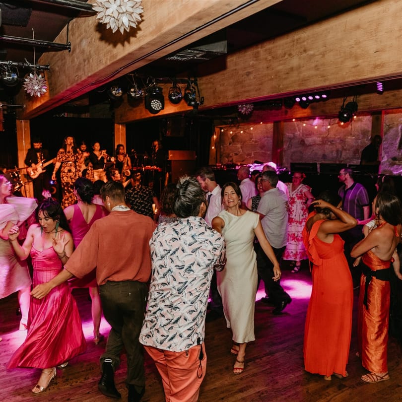 Guests dance on a wooden floor with pink and purple lighting, a band playing in the background.