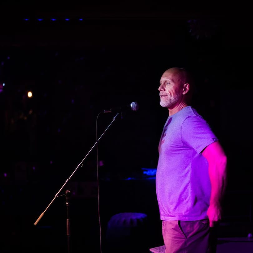 A man with a beard stands on a dark stage, illuminated by purple and blue lights.