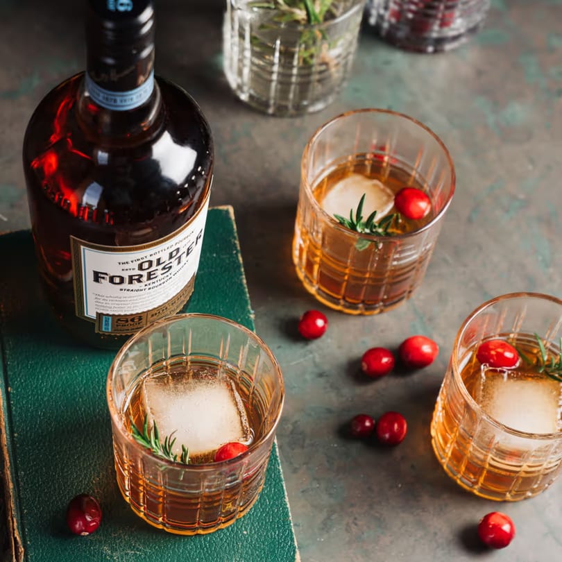 Old Forester bourbon bottle next to two cocktails with ice, cranberries, and rosemary.