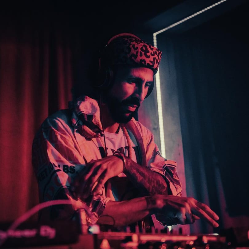 A DJ in a leopard print hat and headphones mixing music under red and blue lights.