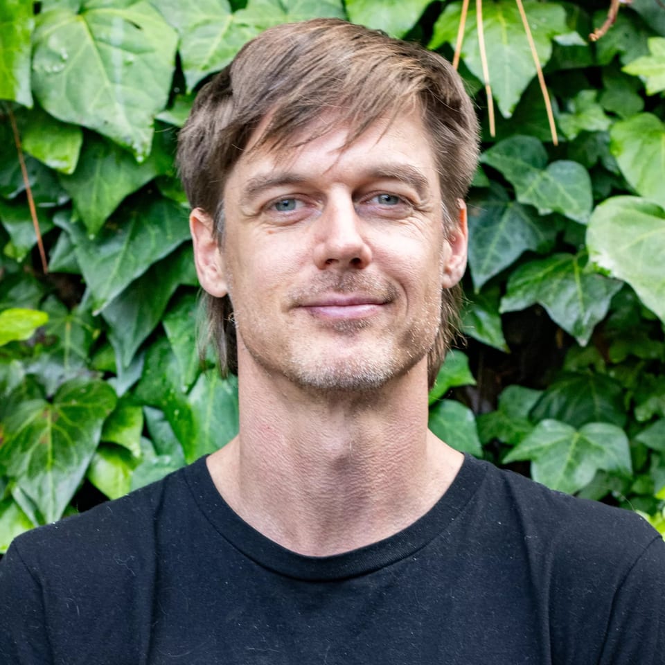 A man with blue eyes and brown hair smiles, standing in front of a green ivy wall.