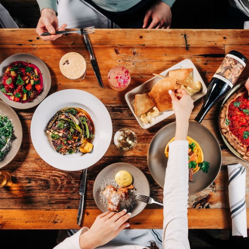 Overhead view of a wooden table laden with various New American dishes, drinks, and hands reaching for food.