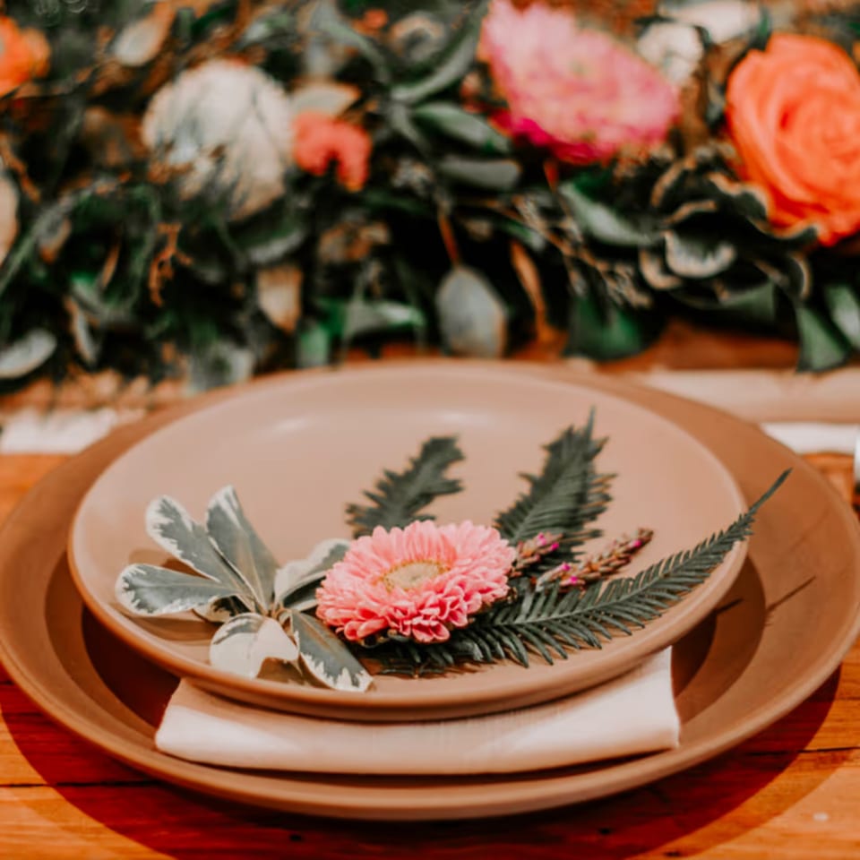 A Stone House place setting with a pink flower, green leaves, and pine sprigs on a plate.