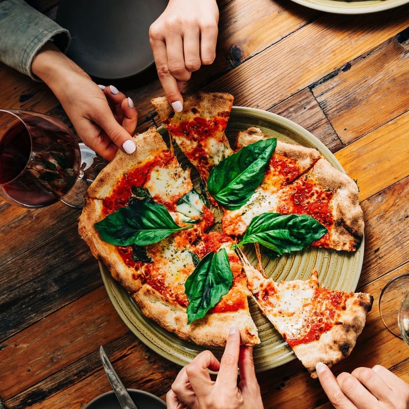 Hands reaching for slices of a Stone House pizza with basil and red wine on a wooden table.